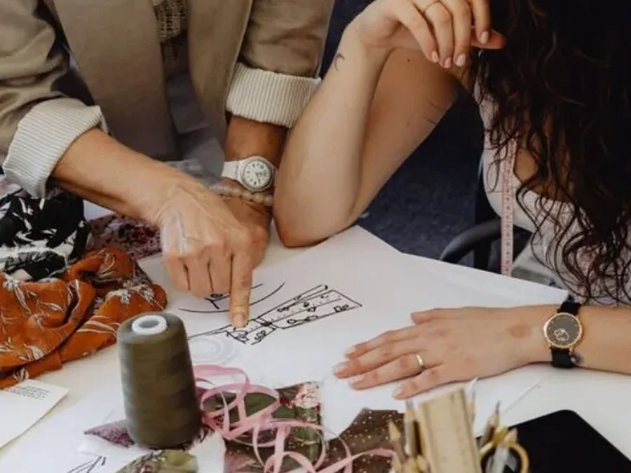 Two people discussing a fashion drawing beside fabric and sewing materials